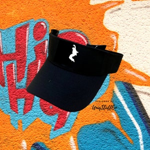May include: Black visor with a white silhouette of a person doing a backflip on the front. The text "DESIGNED BY UniqStuffCo" is printed on the bottom right side of the visor.