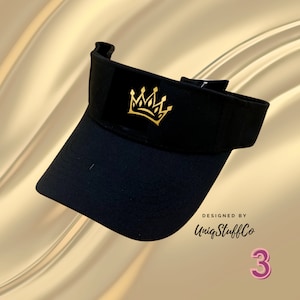 May include: Black visor with a gold crown design. The visor has a black strap and the text "DESIGNED BY UniqStuffCo" is printed on the side.