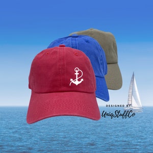 May include: Three cotton baseball caps with an anchor design. The caps are red, blue, and green. The caps are on a blue background with a sailboat in the distance. The text "DESIGNED BY UniqStuffCo" is at the bottom of the image.