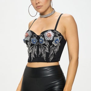 Pode incluir: Um top curto preto com decote coração e bordado floral. O bordado apresenta flores prateadas, azuis e rosas com folhas de contas.