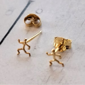 Puede incluir: Pendientes de botón dorados con forma de persona corriendo. Los pendientes son pequeños y delicados, con un diseño sencillo. Se ven un poste y un cierre de pendiente.