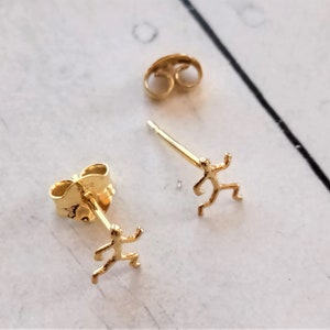 Könnte beinhalten: Goldfarbene Ohrstecker in Form einer rennenden Person. Die Ohrringe haben einen Stecker und einen Butterfly-Verschluss. Ein Ohrring ist fokussiert, der andere Ohrring und der Verschluss sind im Hintergrund sichtbar.