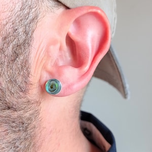 Puede incluir: Primer plano de la oreja de una persona con un pendiente de plata con una concha de abulón redonda con patrones iridiscentes verdes y azules.