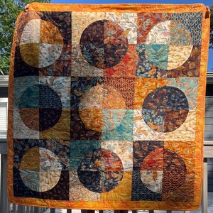 May include: A square quilt featuring a grid of nine large circles set against square blocks. The fabric displays a variety of patterns in shades of orange, gold, teal, brown, and cream, with visible machine quilting stitches throughout the surface.