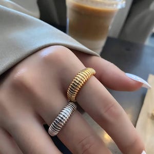 May include: Two ribbed rings, one gold and one silver, are displayed. The rings have a textured, ridged design. The gold ring is on top, and the silver ring is below it. The rings are on a person's finger.