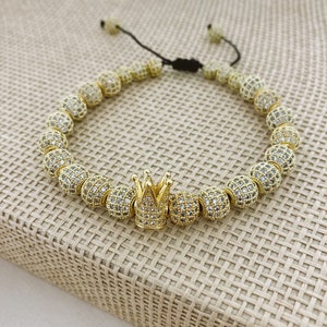 May include: A gold bracelet with a crown charm. The bracelet is made of small, round beads that are covered in clear rhinestones.