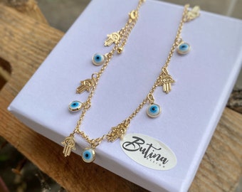 Puede incluir: Collar de cadena dorada con múltiples dijes, incluyendo dijes de ojo azul mal de ojo y mano de hamsa. El collar está sobre un fondo blanco con una etiqueta que dice "Butina Design".