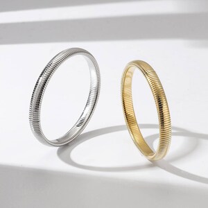 May include: Two bangle bracelets. One is silver-colored, the other is gold-colored. Both bracelets have a ribbed, flexible design. The bracelets are displayed on a white surface, with shadows adding depth.