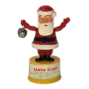 May include: A vintage Santa Claus push-button puppet. The figure is dressed in a red suit with a white beard and hat. It holds a silver bell in one hand. The base is yellow with text that reads "SANTA CLAUS PUSH BUTTON PUPPET".