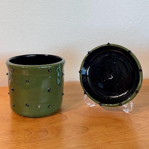 May include: A cylindrical olive green ceramic planter with a glossy black interior and small black spikes protruding from the exterior surface. The planter is accompanied by a matching circular olive green saucer with a black rim and spikes.