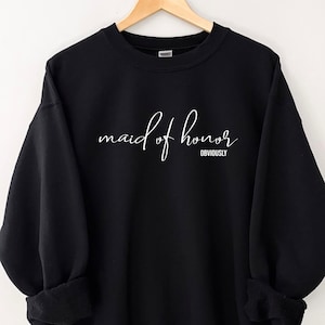 May include: Black sweatshirt with the words "maid of honor" in white script, with "obviously" below. The long-sleeved top is displayed on a wooden hanger.