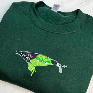 May include: Dark green sweatshirt with an embroidered design. The design features a green character peeking out of a zipper with the words "EW PEOPLE" embroidered on the zipper.