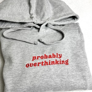 May include: A gray hoodie with a drawstring hood and the embroidered text "probably overthinking" in red. The sweatshirt is folded and lies on a white, textured surface. The hoodie is made of a soft material.