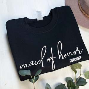 May include: Black crew neck sweatshirt with the words "maid of honor OBVIOUSLY" in white script. A neutral-toned hat and greenery are in the background. The sweatshirt is folded neatly on a white surface.