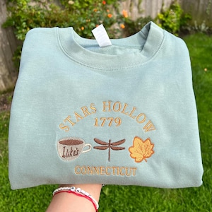 May include: A pale blue sweatshirt featuring embroidered text and images. The design includes "STARS HOLLOW 1779 CONNECTICUT" with a coffee cup, dragonfly, and leaf. The sweatshirt is folded and held up by a person.