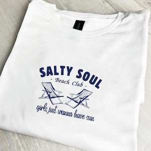 Op de afbeelding: Wit T-shirt met een marineblauwe opdruk. De opdruk toont de tekst "Salty Soul Beach Club", twee strandstoelen en de zin "girls just wanna have sun".