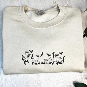May include: A cream-colored sweatshirt featuring a black graphic of cats dressed as ghosts with bats. The crew neck sweatshirt is folded on a light wood surface. The design is centered on the chest.