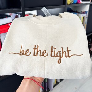 Op de afbeelding: Cr&egrave;mekleurige sweatshirt met de geborduurde tekst "be the light" in bruin. De opgevouwen sweatshirt toont de tekst duidelijk. De stof ziet er zacht en comfortabel uit, ideaal voor dagelijks gebruik.