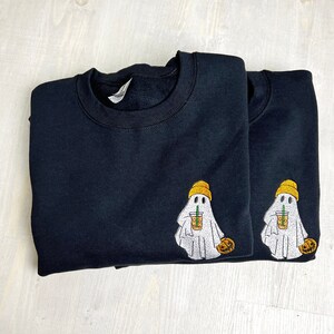 Op de afbeelding: Twee zwarte sweaters met ronde hals en een geborduurd ontwerp van een witte geest met een mosterdgele muts. De geest houdt een doorzichtige plastic beker met een oranje drankje en een groen rietje vast, en in de andere hand een kleine oranje pompoen.