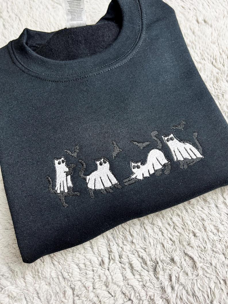 May include: Black sweatshirt featuring an embroidered design of four cats dressed as ghosts with bats above them. The cats are white with black details, and the bats are black. The sweatshirt is made of a soft material.