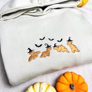 Op de afbeelding: Een lichtbeige hoodie met een capuchon met trekkoord. De voorkant heeft een geborduurd ontwerp van vier dinosaurussen met heksenhoeden, met zwarte vleermuizen die erboven vliegen. Twee pompoenen staan op de voorgrond.