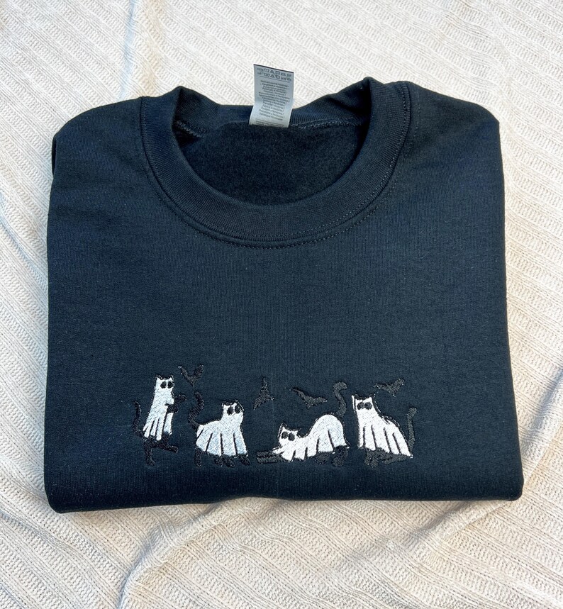 May include: Black crewneck sweatshirt featuring an embroidered design of white ghost cats and black bats. The cats are in various poses. The sweatshirt is folded neatly.