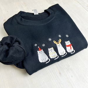 Peut inclure: Sweat-shirt noir avec broderie de chats de No&euml;l. Le motif pr&eacute;sente quatre chats gris avec des accessoires festifs : un bonnet de P&egrave;re No&euml;l, des guirlandes lumineuses, des bois de renne et une &eacute;charpe rouge. Des flocons de neige blancs sont brod&eacute;s au-dessus des chats.