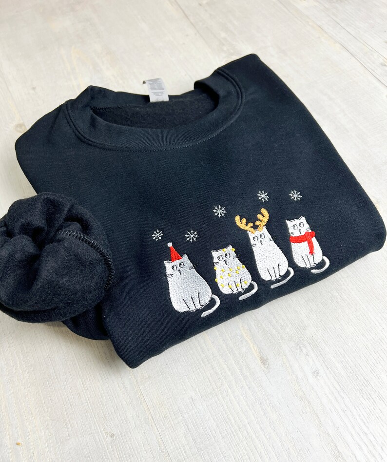Peut inclure: Sweat-shirt noir avec broderie de chats de No&euml;l. Le motif pr&eacute;sente quatre chats gris avec des accessoires festifs : un bonnet de P&egrave;re No&euml;l, des guirlandes lumineuses, des bois de renne et une &eacute;charpe rouge. Des flocons de neige blancs sont brod&eacute;s au-dessus des chats.