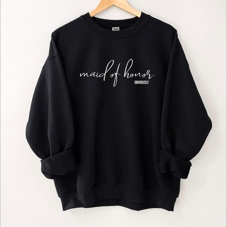 May include: Black sweatshirt with the words "maid of honor" in white script, with "obviously" below. The long-sleeved top is displayed on a wooden hanger.