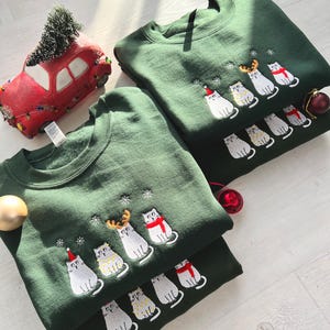 Peut inclure: Deux sweat-shirts vert fonc&eacute; avec des motifs de chats brod&eacute;s. Chaque chat porte un accessoire de f&ecirc;te, comme un chapeau de P&egrave;re No&euml;l ou une &eacute;charpe. Une voiture rouge avec un sapin de No&euml;l et des ornements sont &eacute;galement pr&eacute;sents.