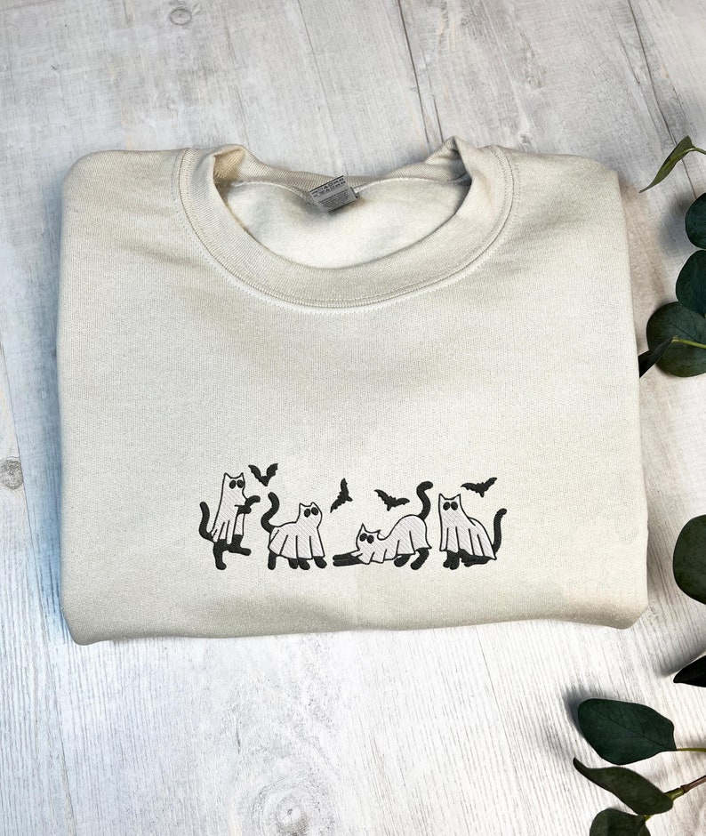 May include: A cream-colored sweatshirt featuring a black graphic of cats dressed as ghosts with bats. The crew neck sweatshirt is folded on a light wood surface. The design is centered on the chest.