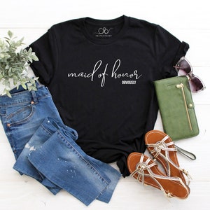 May include: Black t-shirt with the words "maid of honor" in cursive and the word "obviously" below. The shirt is styled with blue jeans, brown sandals, a green wallet, and sunglasses. A sprig of greenery is also included.