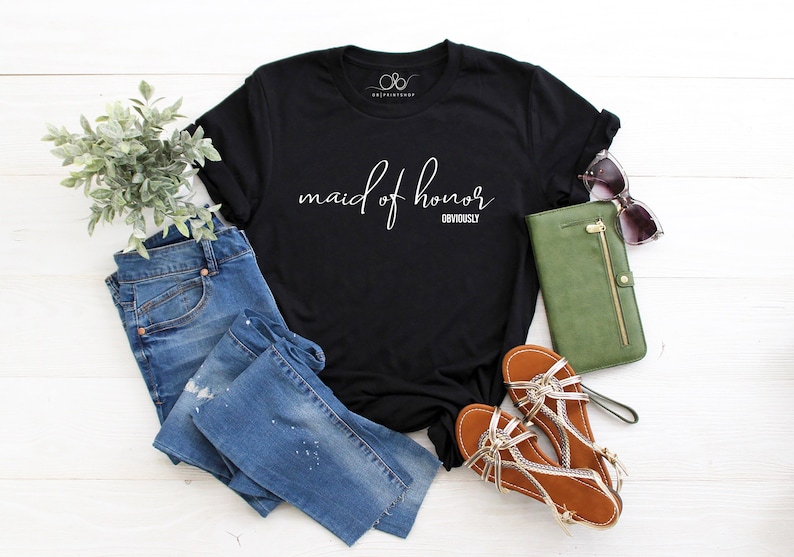 May include: Black t-shirt with the words "maid of honor" in cursive and the word "obviously" below. The shirt is styled with blue jeans, brown sandals, a green wallet, and sunglasses. A sprig of greenery is also included.
