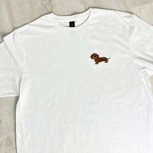 May include: White t-shirt with a small embroidered brown dachshund dog on the chest. The shirt has a crew neck and short sleeves. The dachshund is detailed with brown and tan colors.