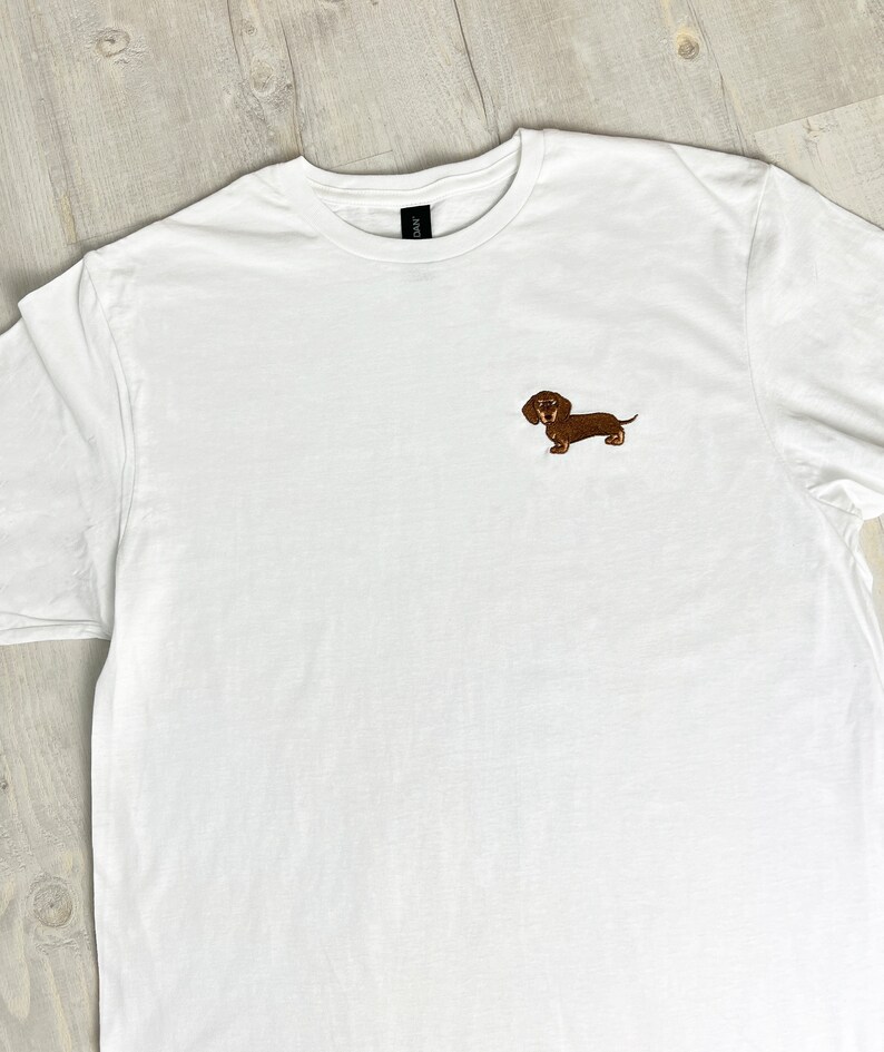 May include: White t-shirt with a small embroidered brown dachshund dog on the chest. The shirt has a crew neck and short sleeves. The dachshund is detailed with brown and tan colors.