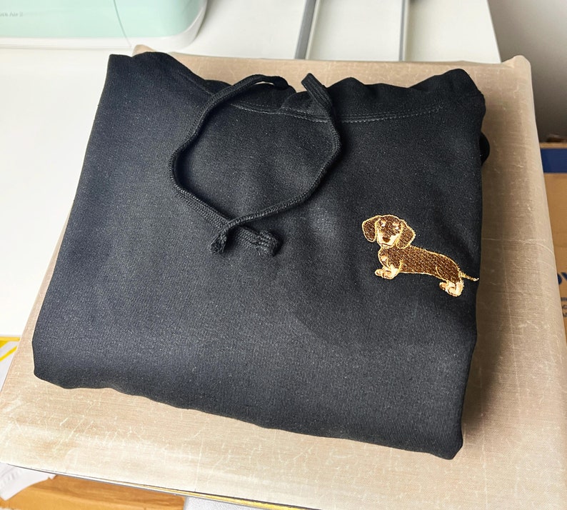 May include: Black hoodie with a drawstring and a gold embroidered dachshund dog on the front. The hoodie is folded neatly on a light brown surface. The dog embroidery is detailed with brown and tan colors.
