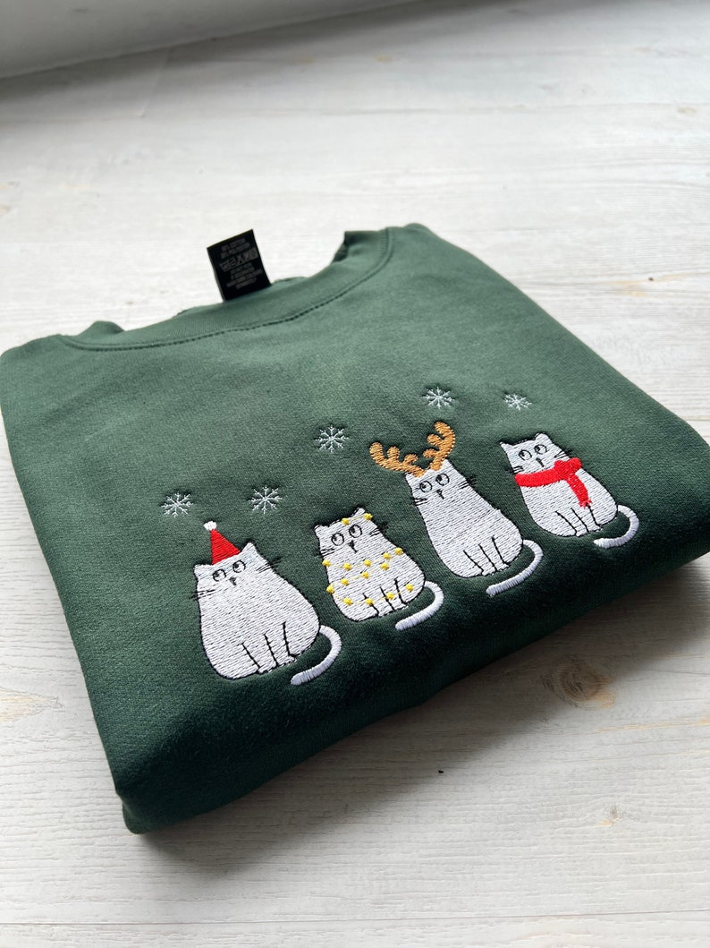 Peut inclure: Un sweat-shirt vert fonc&eacute; avec une broderie de quatre chats blancs. Chaque chat est orn&eacute; d'accessoires de vacances festifs, dont un bonnet de P&egrave;re No&euml;l, des guirlandes lumineuses, des bois de renne et une &eacute;charpe rouge. Des flocons de neige sont brod&eacute;s au-dessus des chats.