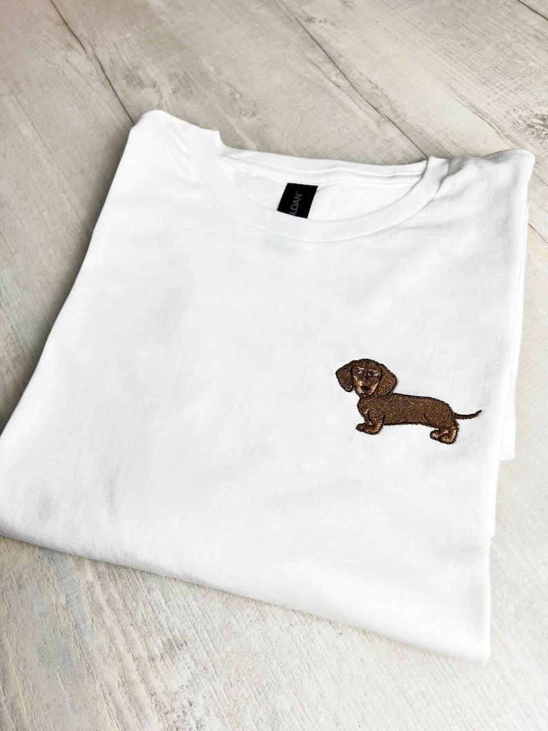 May include: White t-shirt with a brown embroidered dachshund dog on the front. The shirt is folded and lies on a light wood surface. The dog is detailed with brown and black thread.