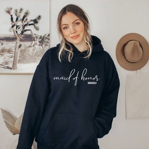 May include: A navy blue hoodie featuring the text "maid of honor" in white cursive lettering. Beneath, the word "obviously" is printed. The sweatshirt has a front pocket and a drawstring hood, perfect for casual wear.