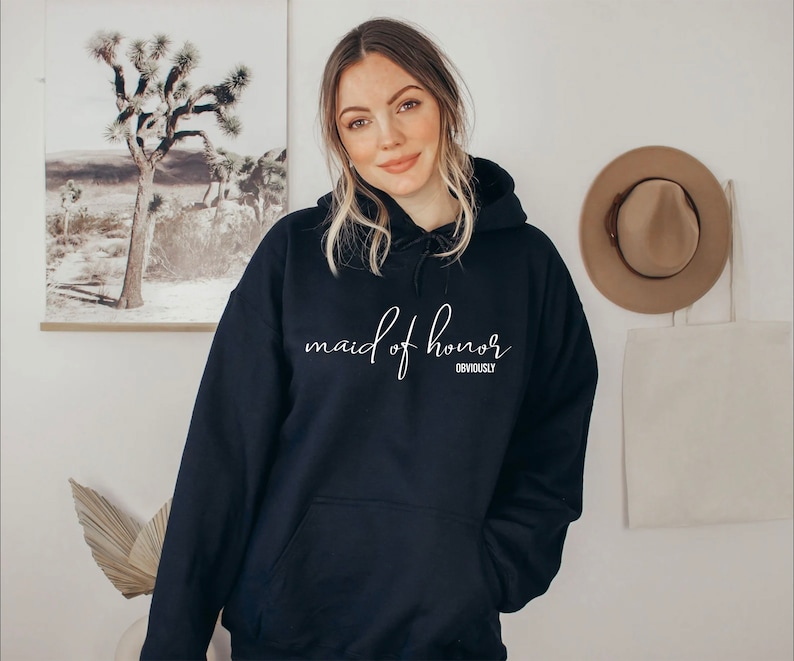 May include: A navy blue hoodie featuring the text "maid of honor" in white cursive lettering. Beneath, the word "obviously" is printed. The sweatshirt has a front pocket and a drawstring hood, perfect for casual wear.