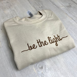 Op de afbeelding: Een beige sweatshirt met de geborduurde zin "be the light" in bruin schrift. De sweatshirt is netjes opgevouwen op een licht oppervlak met houtnerf. Een label is zichtbaar bij de kraag.