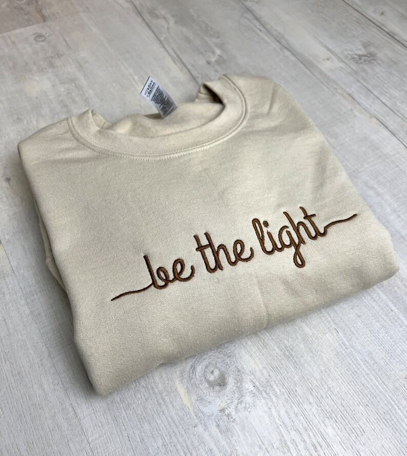 Op de afbeelding: Een beige sweatshirt met de geborduurde zin "be the light" in bruin schrift. De sweatshirt is netjes opgevouwen op een licht oppervlak met houtnerf. Een label is zichtbaar bij de kraag.