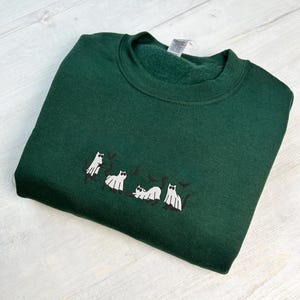 May include: A dark green sweatshirt with a crew neck. The sweatshirt features an embroidered design of white ghost cats with black accents. The cats are in various poses, creating a playful Halloween theme.