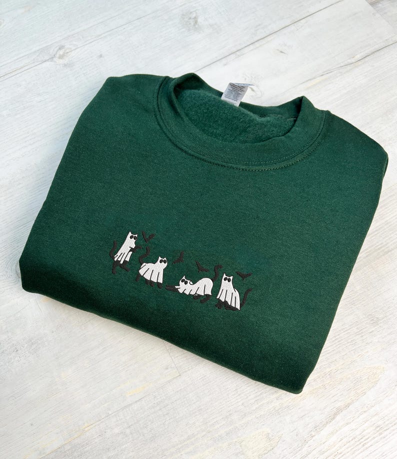 May include: A dark green sweatshirt with a crew neck. The sweatshirt features an embroidered design of white ghost cats with black accents. The cats are in various poses, creating a playful Halloween theme.