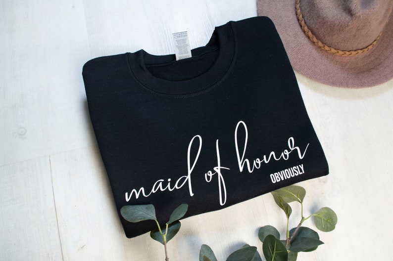 May include: Black crew neck sweatshirt with the words "maid of honor OBVIOUSLY" in white script. A neutral-toned hat and greenery are in the background. The sweatshirt is folded neatly on a white surface.