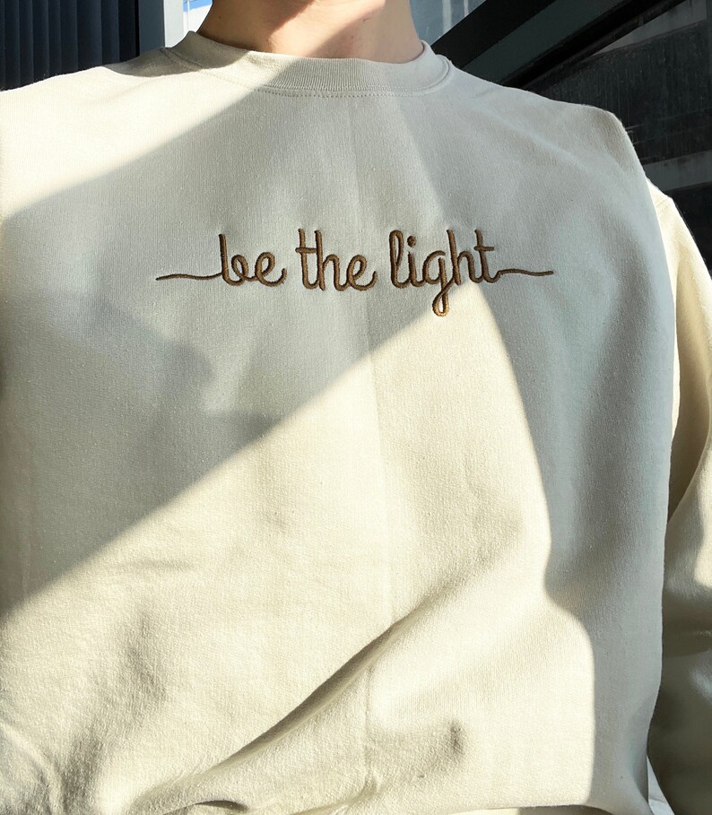 Op de afbeelding: Cr&egrave;mekleurige sweatshirt met de geborduurde tekst "be the light" in bruin. De crew neck sweatshirt is gemaakt van een zacht materiaal. De afbeelding is goed belicht, waardoor de textuur van de stof en de geborduurde tekst zichtbaar zijn.