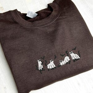 May include: A dark brown sweatshirt with a crew neck. The sweatshirt features an embroidered design of cats dressed as ghosts with bats. The cats are in various poses, creating a Halloween theme.