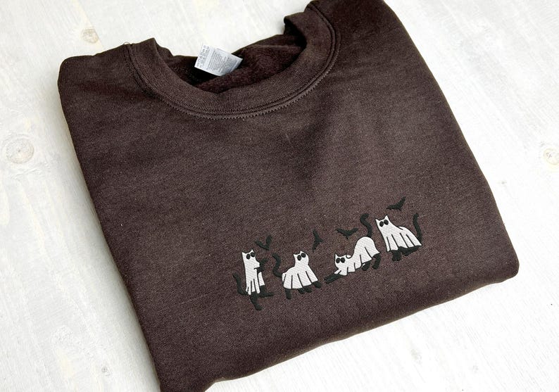 May include: A dark brown sweatshirt with a crew neck. The sweatshirt features an embroidered design of cats dressed as ghosts with bats. The cats are in various poses, creating a Halloween theme.