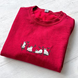 May include: A folded red sweatshirt with a crew neck. The sweatshirt features an embroidered design of white cats with black accents. The cats are in various poses, with some holding hearts.