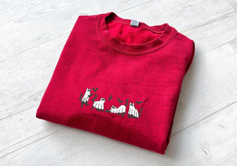 May include: A folded red sweatshirt with a crew neck. The sweatshirt features an embroidered design of white cats with black accents. The cats are in various poses, with some holding hearts.